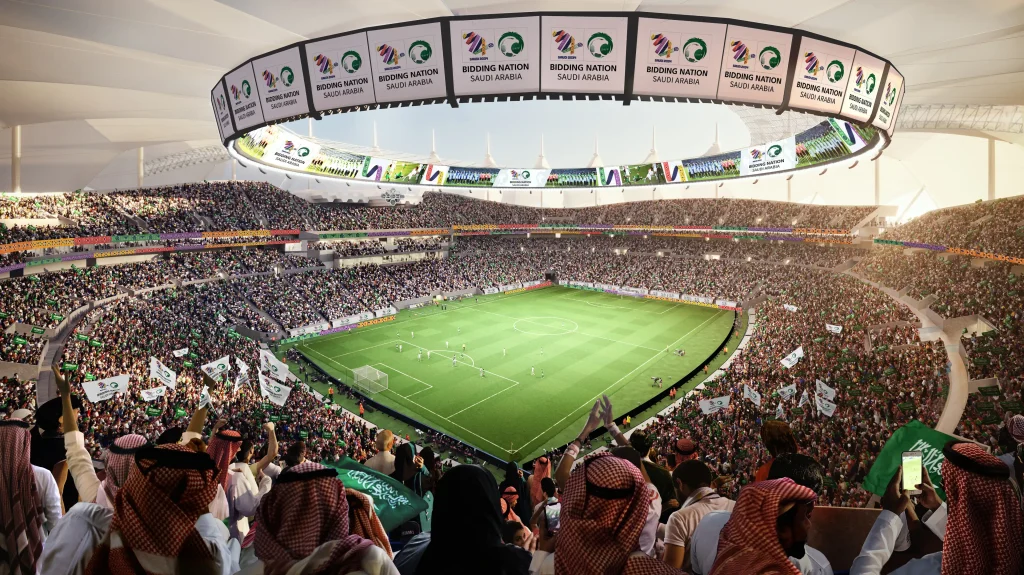 Packed football stadium in Saudi Arabia showing thousands of enthusiastic fans watching a match, with 'Bidding Nation Saudi Arabia' banners displayed on the circular roof structure above the pitch